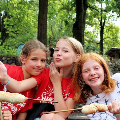 Drei lächelnde Mädchen mit einem gegrillten Maiskolben im Sommercamp Heino.