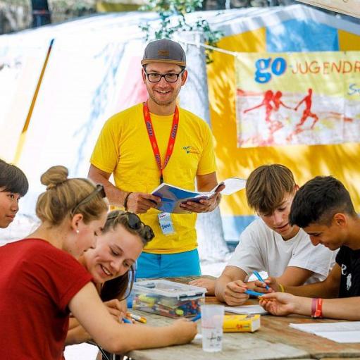 Fröhliche Gruppe von Jugendlichen bei einer Lern- und Freizeitaktivität in einem Sommercamp, betreut von einem Betreuer in gelbem Shirt.
